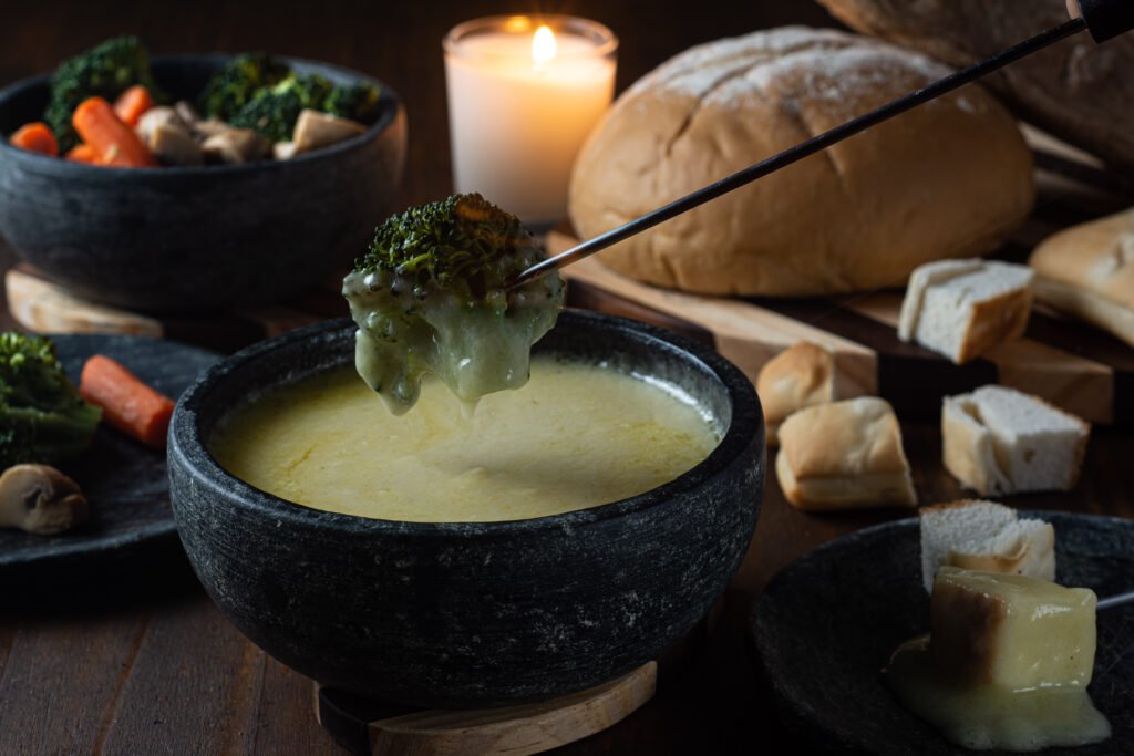 Em destaque tem um pequeno brocolis sendo mergulhado no fondue de queijo. Ao fundo da foto, tem pães e legumes que podem acompanhar o fondue