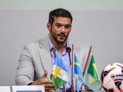 Samir Xaud em evento da CBF, vestindo terno e gravata, com bandeiras ao fundo
