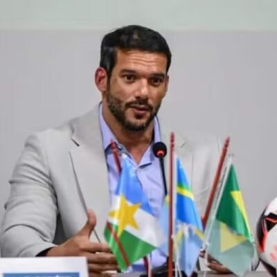 Samir Xaud em evento da CBF, vestindo terno e gravata, com bandeiras ao fundo