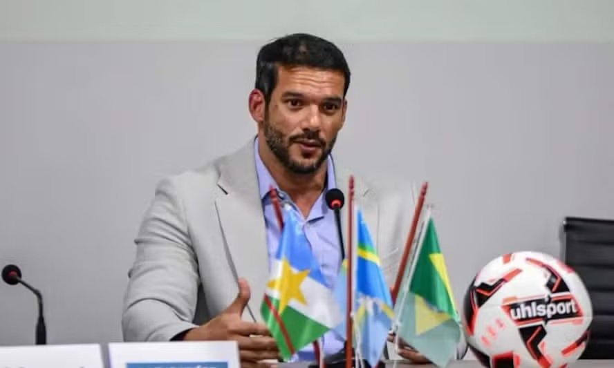 Samir Xaud em evento da CBF, vestindo terno e gravata, com bandeiras ao fundo