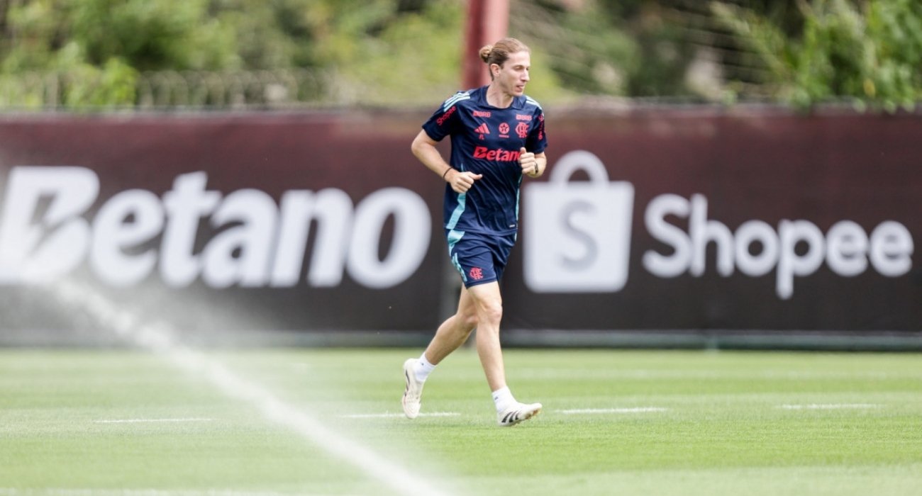 Preparação do Flamengo em meio aos desfalques para o jogo contra o Fortaleza Filipe Luís, técnico do Flamengo, correndo no gramado do centro de treinamento, com um sistema de irrigação em primeiro plano. Ao fundo, banners com os patrocinadores Betano e Shopee são visíveis. A imagem reflete a importância da preparação física em meio aos desafios do time.