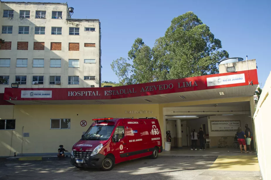 Entrada do Hospital Estadual Azevedo Lima com ambulância vermelha estacionada na frente, pessoas aguardando na entrada e céu claro ao fundo