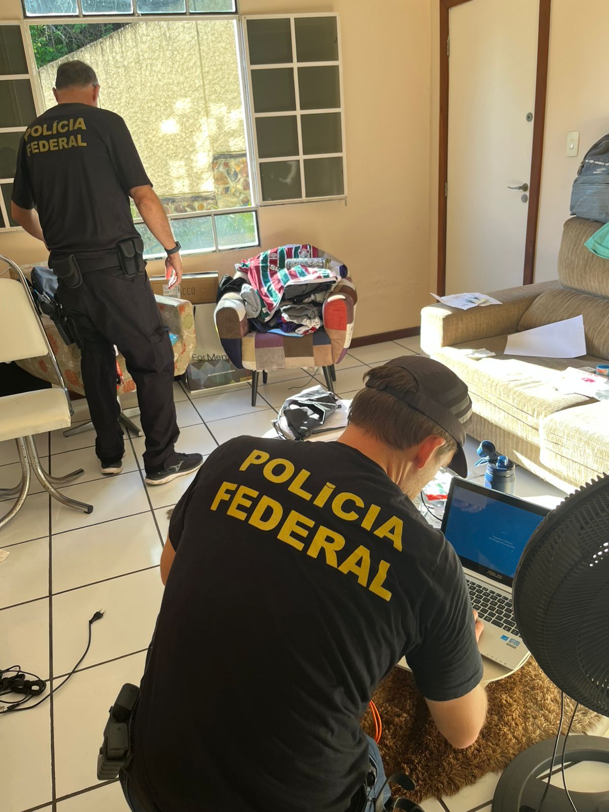 Policiais federais analisam documentos e equipamentos dentro de residência alvo da operação contra fraude.
