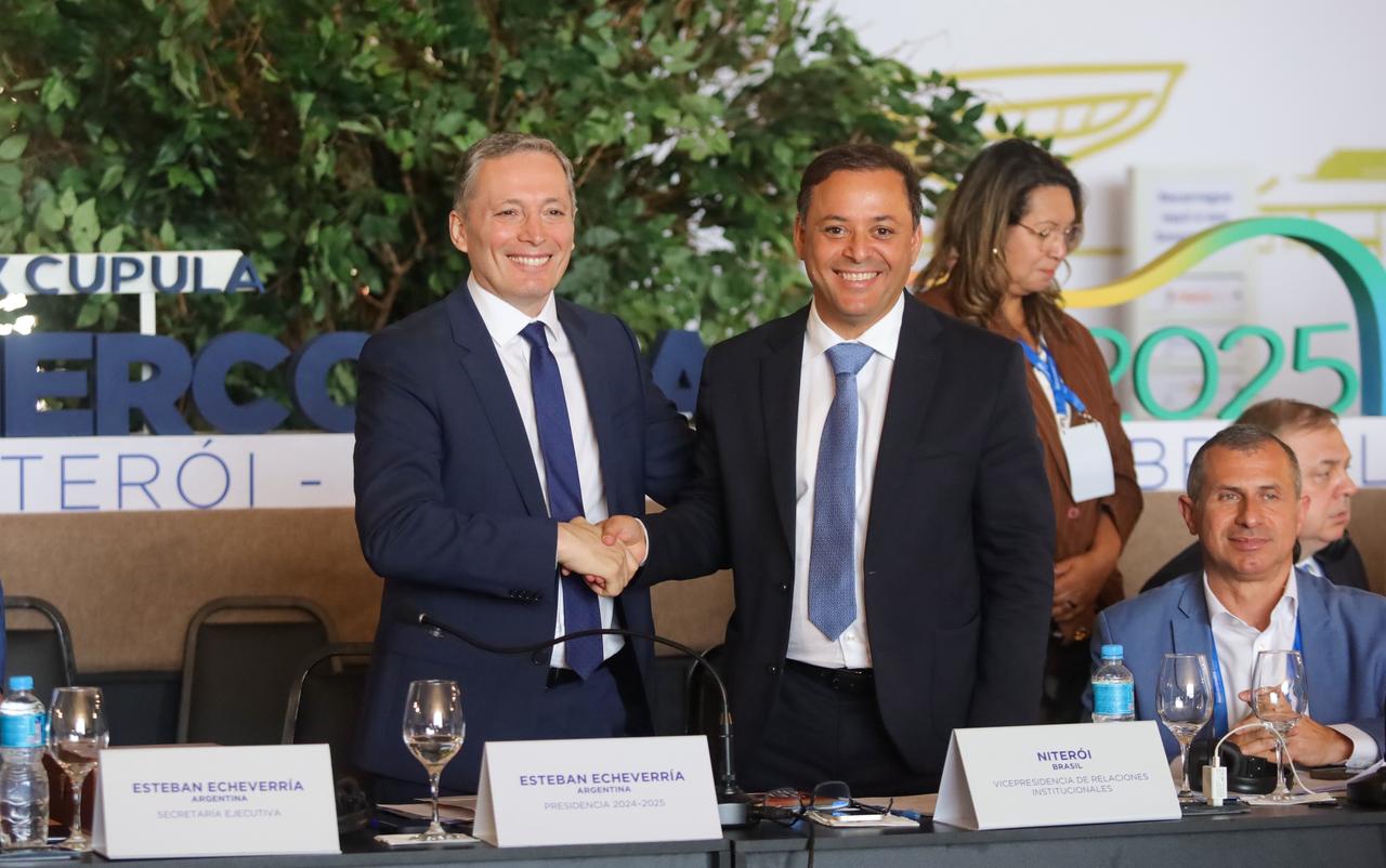 Esteban Echeverría e Rodrigo Neves, vestidos de terno e gravata, apertam as mãos sorrindo. Ao fundo, um banner com a inscrição "XXX Cúpula Mercocidades Niterói 2025 Brasil".