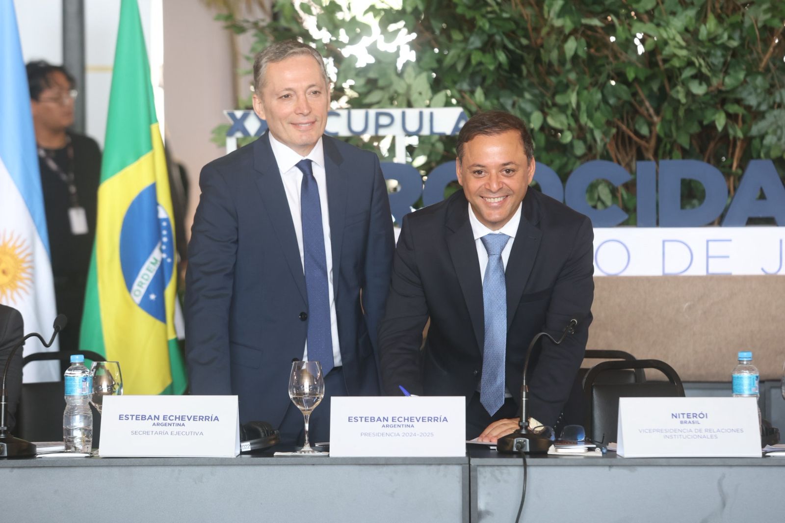 Rodrigo Neves, sorrindo, inclina-se para assinar um documento sobre a mesa. Ao seu lado, Esteban Echeverría observa. Bandeiras do Brasil e da Argentina aparecem ao fundo.