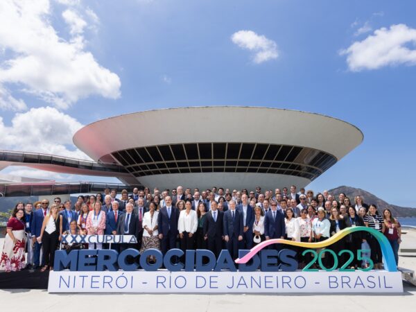 Grande grupo de dezenas de autoridades posa para fotografia nas escadarias externas do MAC Niterói, sob céu azul. Em primeiro plano, um letreiro diz "Mercocidades 2025 Niterói".