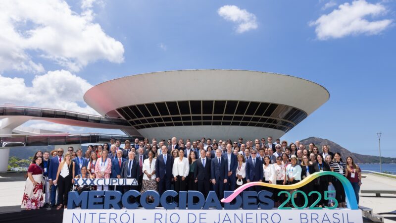 Grande grupo de dezenas de autoridades posa para fotografia nas escadarias externas do MAC Niterói, sob céu azul. Em primeiro plano, um letreiro diz "Mercocidades 2025 Niterói".
