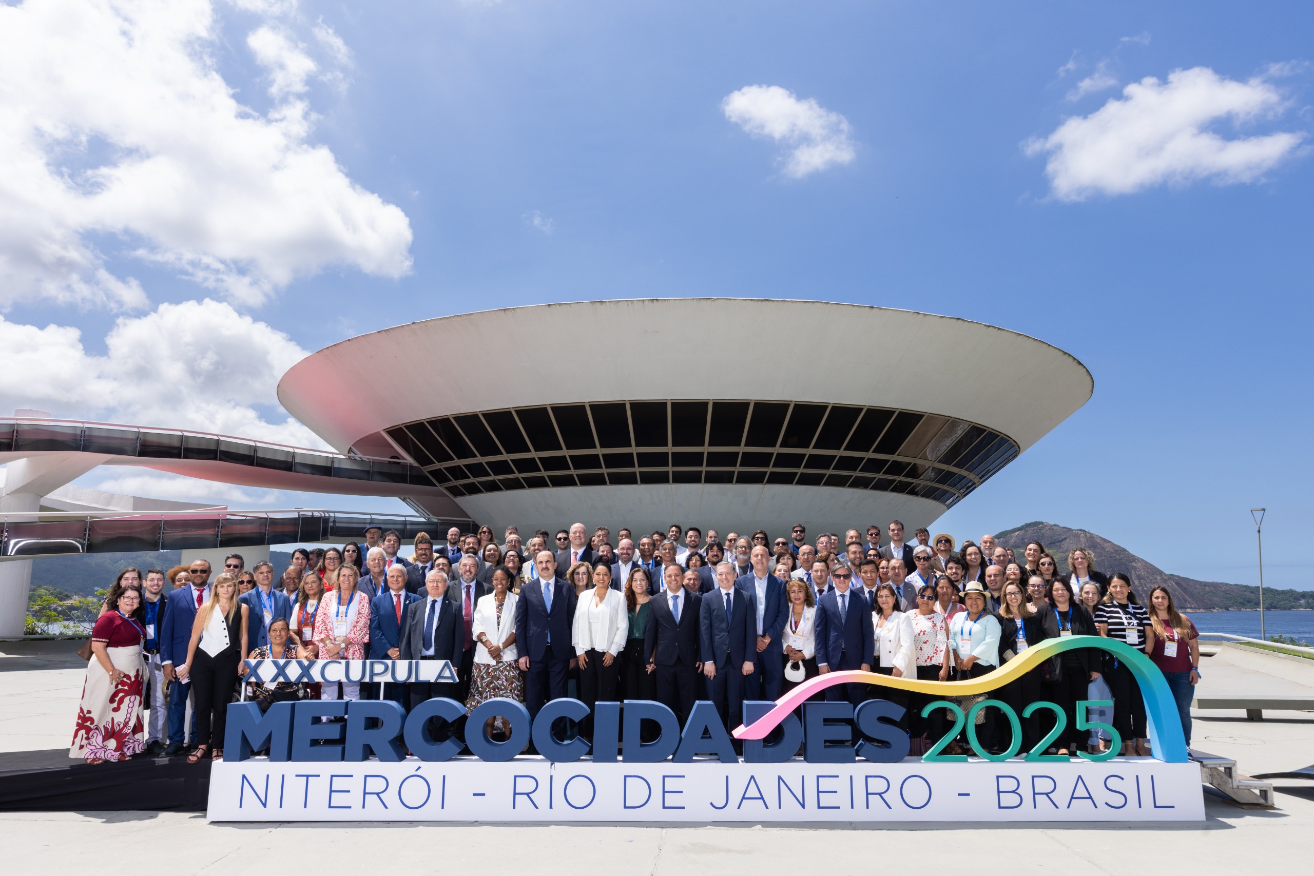 Grande grupo de dezenas de autoridades posa para fotografia nas escadarias externas do MAC Niterói, sob céu azul. Em primeiro plano, um letreiro diz "Mercocidades 2025 Niterói".