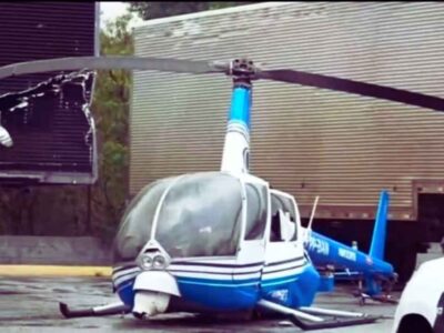 Helicóptero da Band, modelo Robinson R44 azul e branco, pousado em pátio de empresa com danos na estrutura após pane.