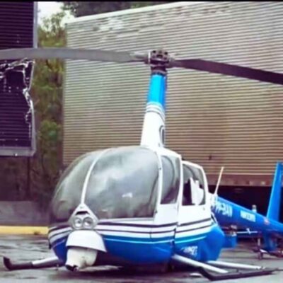 Helicóptero da Band, modelo Robinson R44 azul e branco, pousado em pátio de empresa com danos na estrutura após pane.