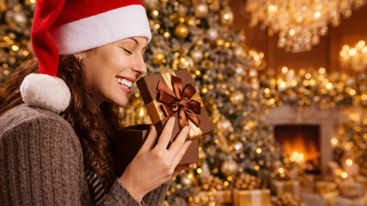 Mulher sorrindo com chapéu de natal ao abrir um presente sofisticado, simbolizando os ganhos e mimos de uma baby sugar por ano.