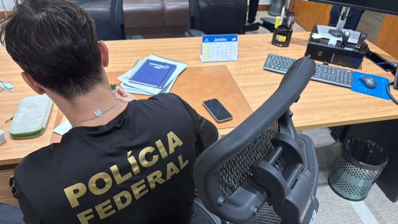 Agente da Polícia Federal de costas, sentado à mesa com documentos e computador, durante operação no Rioprevidência.