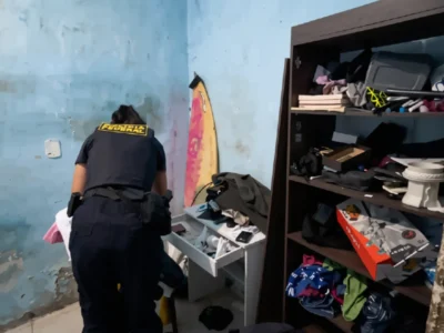Agente da Polícia Federal de costas, utilizando colete tático escuro com a inscrição "FEDERAL" em letras amarelas, inspeciona gavetas de um móvel branco durante busca domiciliar.