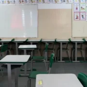 Imagem de uma sala de aula vazia, na imagem tem cadeiras escolares e dois quadros na parede para o professor escrever