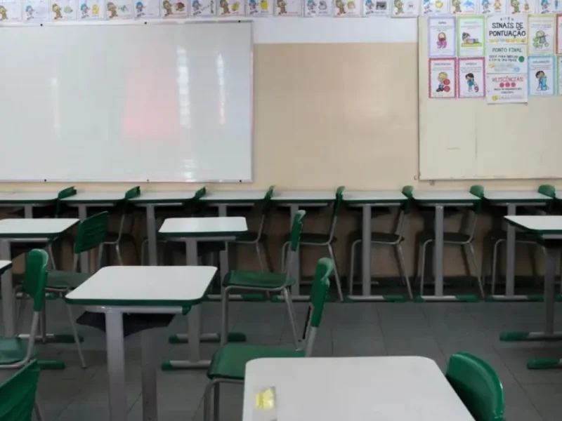 Imagem de uma sala de aula vazia, na imagem tem cadeiras escolares e dois quadros na parede para o professor escrever