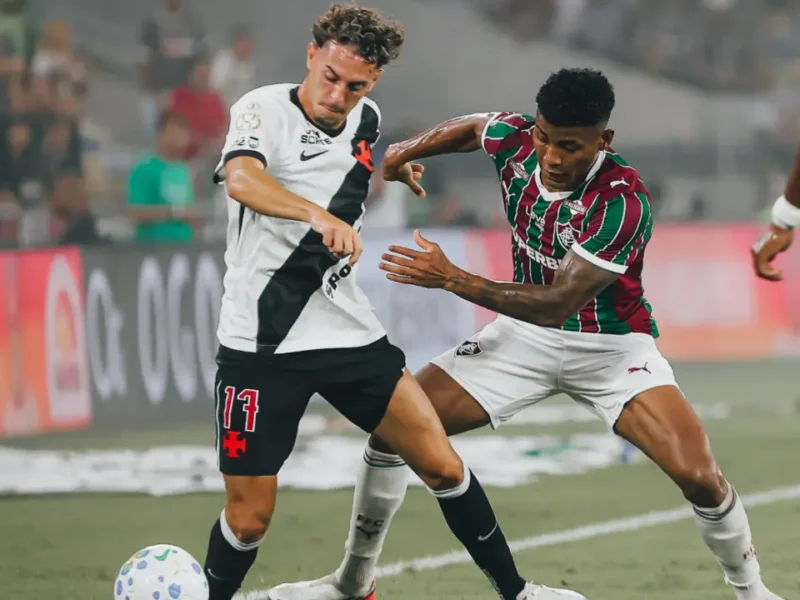 Jogador do Vasco, vestindo uniforme branco com faixa diagonal preta, e atleta do Fluminense, de camisa tricolor, disputam a posse da bola no gramado. A imagem retrata um lance de marcação do confronto Vasco x Fluminense Copa do Brasil.
