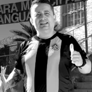 Foto em preto e branco do vereador Sandrinho do Som, de Tanguá (RJ), executado a tiros uma noite após a formatira da filha, em frente à Câmara Municipal, com a placa da instituição ao fundo, segurando papéis e fazendo um sinal de 'positivo'.