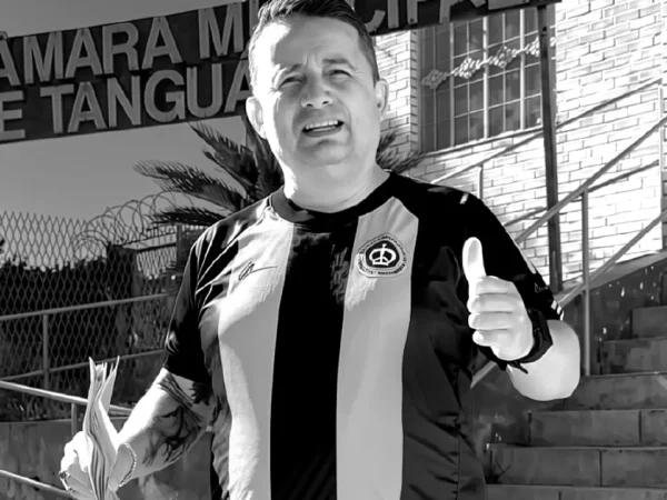 Foto em preto e branco do vereador Sandrinho do Som, de Tanguá (RJ), executado a tiros uma noite após a formatira da filha, em frente à Câmara Municipal, com a placa da instituição ao fundo, segurando papéis e fazendo um sinal de 'positivo'.