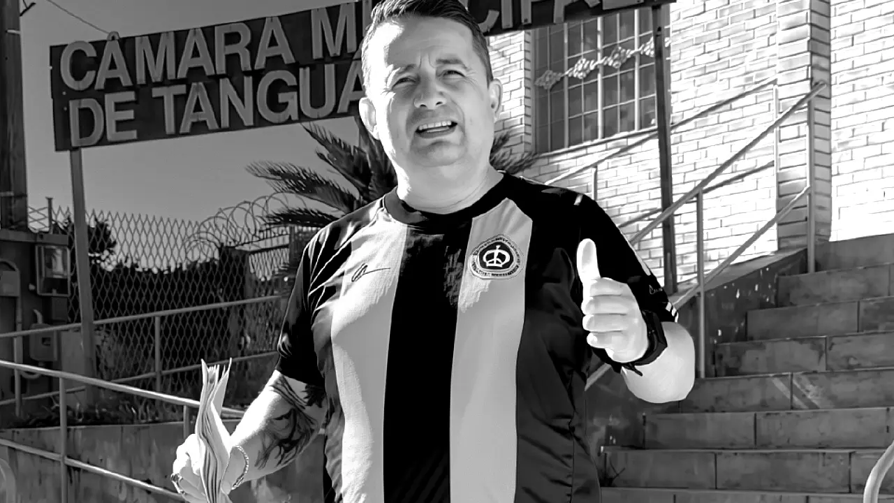 Foto em preto e branco do vereador Sandrinho do Som, de Tanguá (RJ), executado a tiros uma noite após a formatira da filha, em frente à Câmara Municipal, com a placa da instituição ao fundo, segurando papéis e fazendo um sinal de 'positivo'.