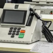 Urna eletrônica ao lado de cola eleitoral em papel com espaços para anotar os números dos candidatos nas Eleições 2026