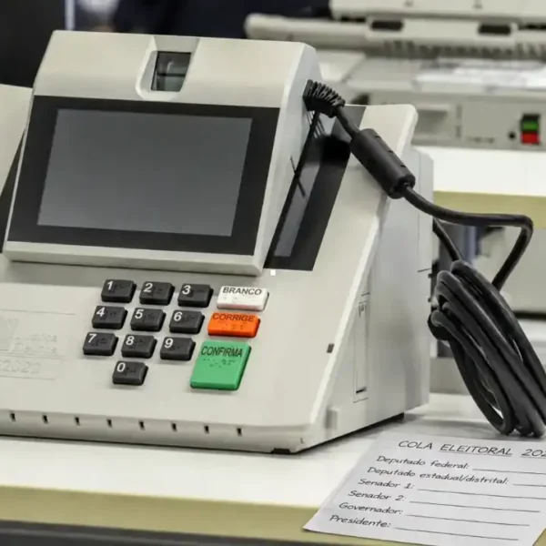 Urna eletrônica ao lado de cola eleitoral em papel com espaços para anotar os números dos candidatos nas Eleições 2026