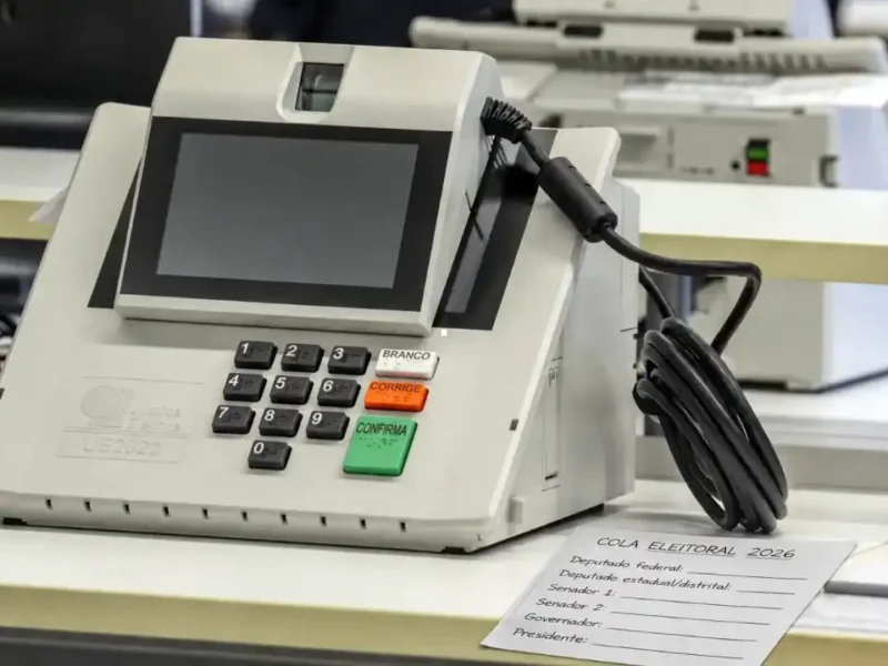 Urna eletrônica ao lado de cola eleitoral em papel com espaços para anotar os números dos candidatos nas Eleições 2026