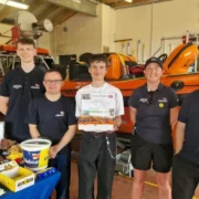 Jovem segura apitos impressos em 3D ao lado de integrantes da RNLI diante de embarcações de resgate.