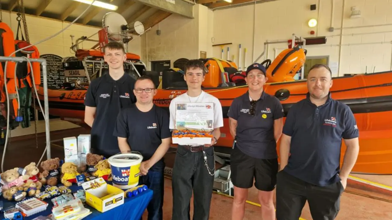 Jovem segura apitos impressos em 3D ao lado de integrantes da RNLI diante de embarcações de resgate. Jovem segura apitos impressos em 3D ao lado de integrantes da RNLI diante de embarcações de resgate.