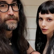 Casal de cabelos longos posa junto diante de uma parede clara