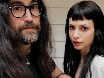 Casal de cabelos longos posa junto diante de uma parede clara