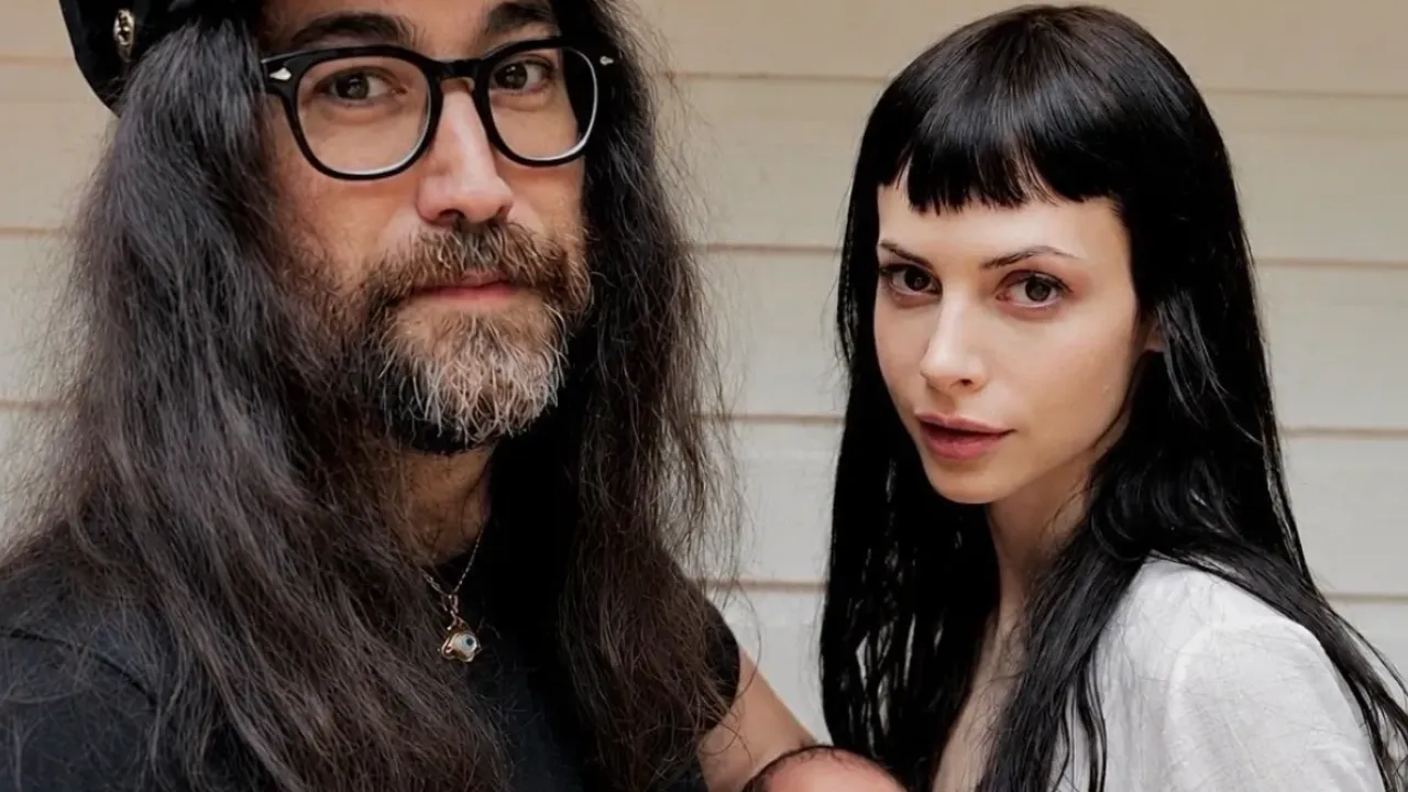 Casal de cabelos longos posa junto diante de uma parede clara