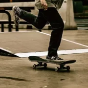 Detalhe das pernas de um skatista executando manobra em uma pista de skate em Niterói, vestindo calça preta e tênis estampado, com um pé sobre a prancha e o outro no ar, tendo uma rampa desfocada ao fundo
