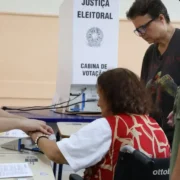 Eleitora em cadeira de rodas recebe atendimento de mesário em seção eleitoral, acompanhada por duas mulheres.