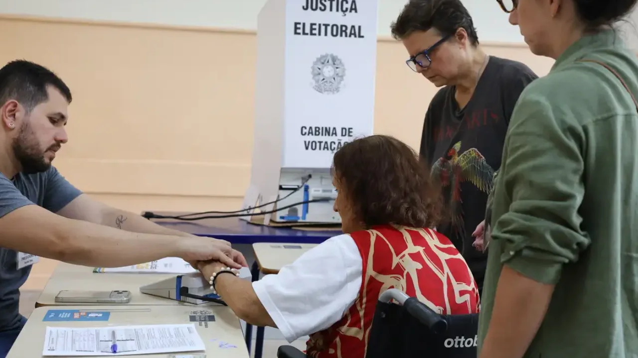 Eleitora em cadeira de rodas recebe atendimento de mesário em seção eleitoral, acompanhada por duas mulheres.