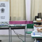 Eleitor utiliza cabine de votação com urna eletrônica enquanto mesários trabalham em uma seção eleitoral.