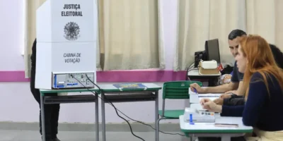Eleitor utiliza cabine de votação com urna eletrônica enquanto mesários trabalham em uma seção eleitoral.