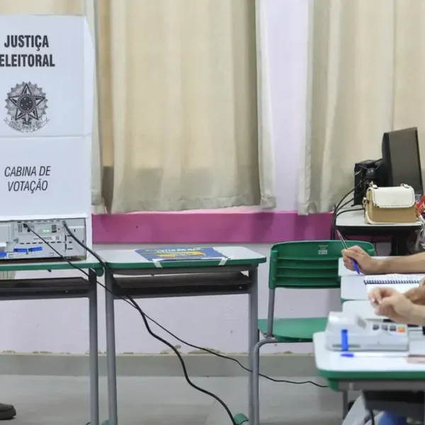 Eleitor utiliza cabine de votação com urna eletrônica enquanto mesários trabalham em uma seção eleitoral.