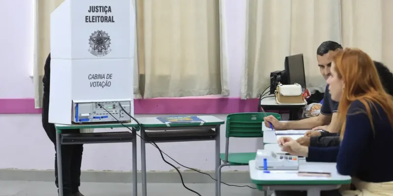 Eleitor utiliza cabine de votação com urna eletrônica enquanto mesários trabalham em uma seção eleitoral.
