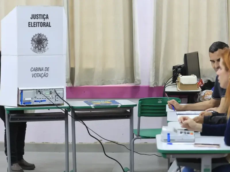 Eleitor utiliza cabine de votação com urna eletrônica enquanto mesários trabalham em uma seção eleitoral.