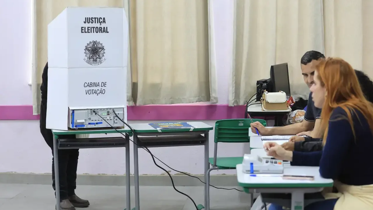 Eleitor utiliza cabine de votação com urna eletrônica enquanto mesários trabalham em uma seção eleitoral.