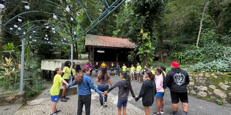Crianças do Clin Social participam de brincadeira coletiva de mãos dadas, em espaço arborizado durante atividade recreativa em Niterói.