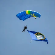 Paraquedista do Exército Brasileiro desce com paraquedas azul, verde e amarelo, transportando uma bandeira durante demonstração aeroterrestre.