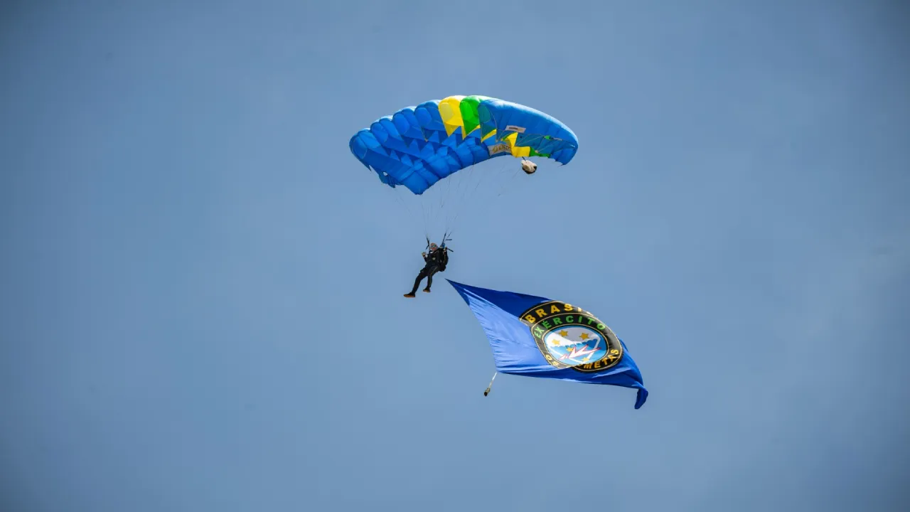 Paraquedista do Exército Brasileiro desce com paraquedas azul, verde e amarelo, transportando uma bandeira durante demonstração aeroterrestre.