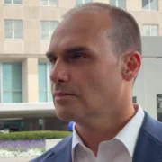 Homem de blazer azul e camisa branca, fotografado de perfil em ambiente externo, diante de um edifício com fachada de vidro.