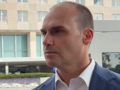 Homem de blazer azul e camisa branca, fotografado de perfil em ambiente externo, diante de um edifício com fachada de vidro.
