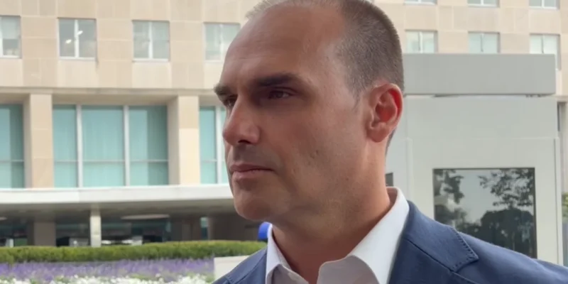 Homem de blazer azul e camisa branca, fotografado de perfil em ambiente externo, diante de um edifício com fachada de vidro.