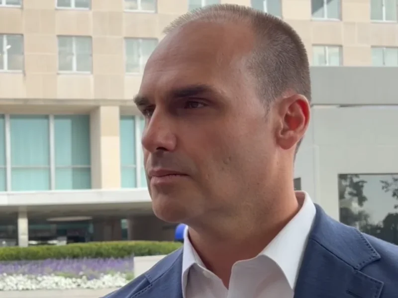 Homem de blazer azul e camisa branca, fotografado de perfil em ambiente externo, diante de um edifício com fachada de vidro.