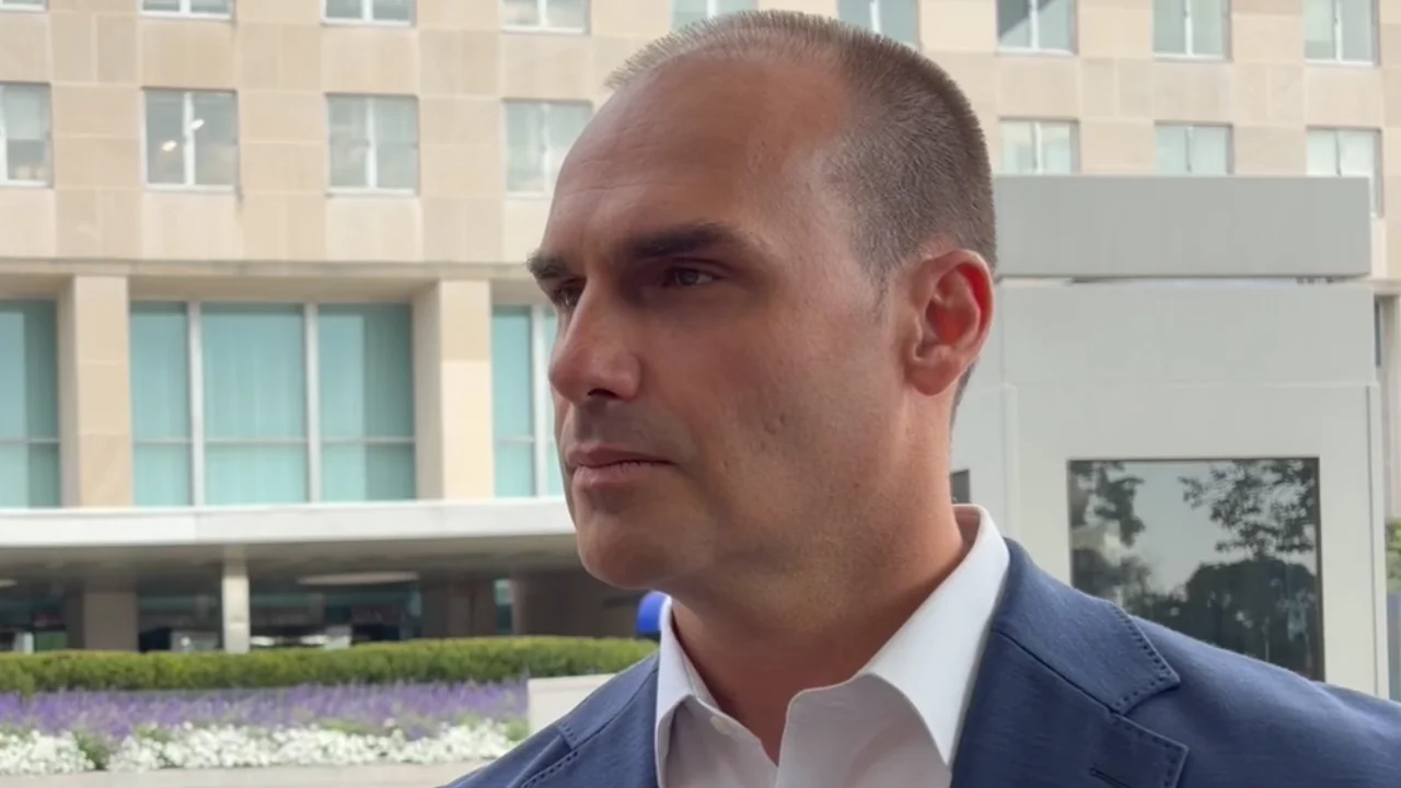 Homem de blazer azul e camisa branca, fotografado de perfil em ambiente externo, diante de um edifício com fachada de vidro.