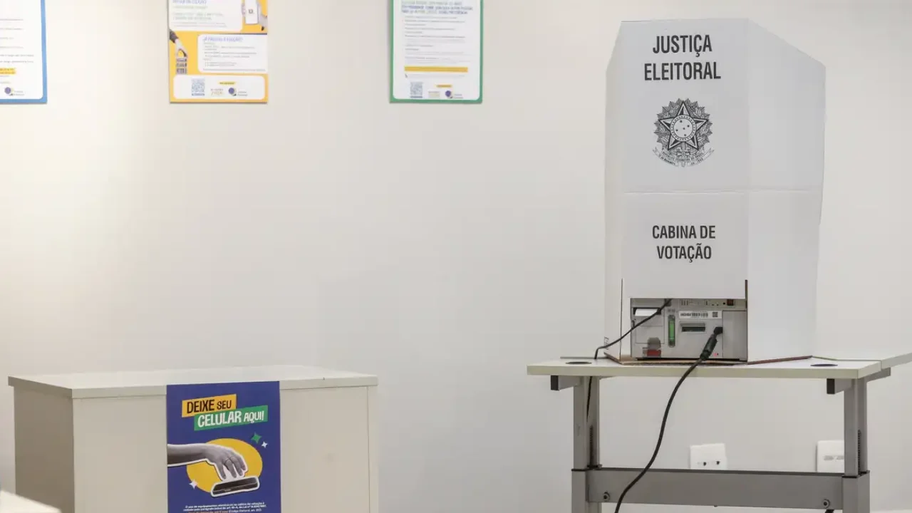 Urna eletrônica instalada em cabine de votação da Justiça Eleitoral, ao lado de compartimento para guardar celulares.