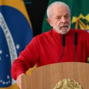 Presidente Lula, de camisa vermelha, discursa diante de dois microfones, com a bandeira brasileira e a bandeira presidencial ao fundo.